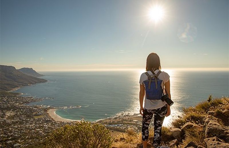 A casually-dressed woman, with a backpack and a Canon EOS RP camera over one shoulder, looks out to sea at a sunset.