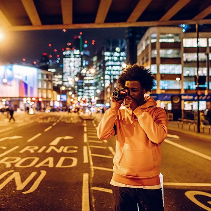 A man stands under a city centre bridge beside a road to take a photo with a Canon camera.