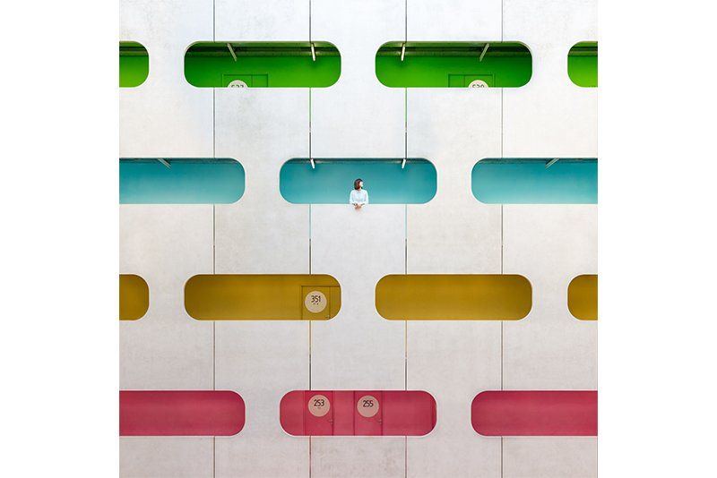 A woman leans out of a blue window in a multi-story atrium with colourful windows. Photo by Daniel Rueda and Anna Devís.