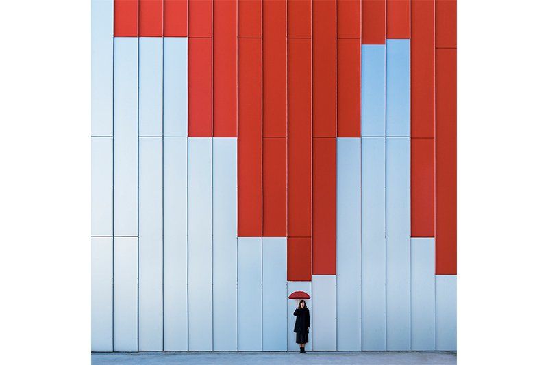 A woman with a red umbrella stands in front of a tall red and blue wall five times taller than her. Photo by Daniel Rueda and Anna Devís.