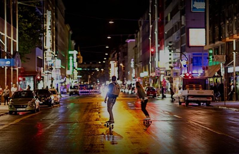 Two people skateboard down a city street at night, lights from buildings reflecting in the wet road surface.