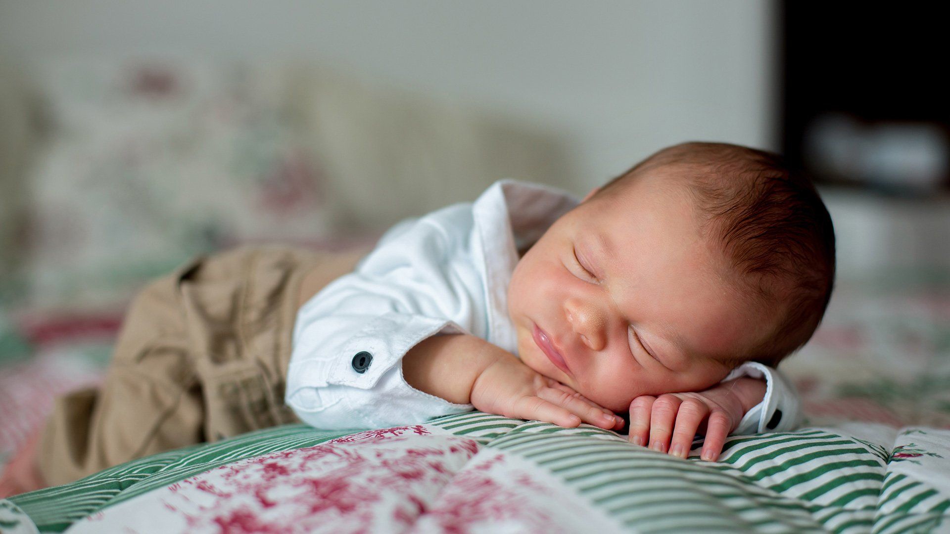 A baby wears a shirt and lies on its front on a green, pink and white blanket.