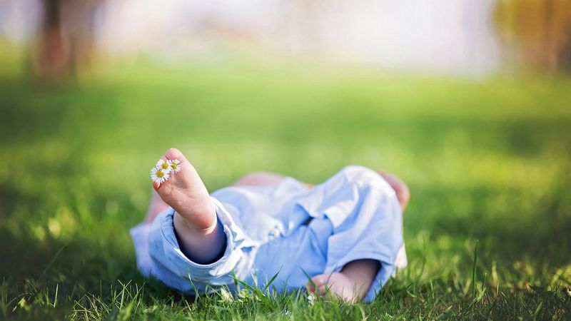 A baby lies on its back in the grass. We just see the bottom of its feet, with daisies between its toes.