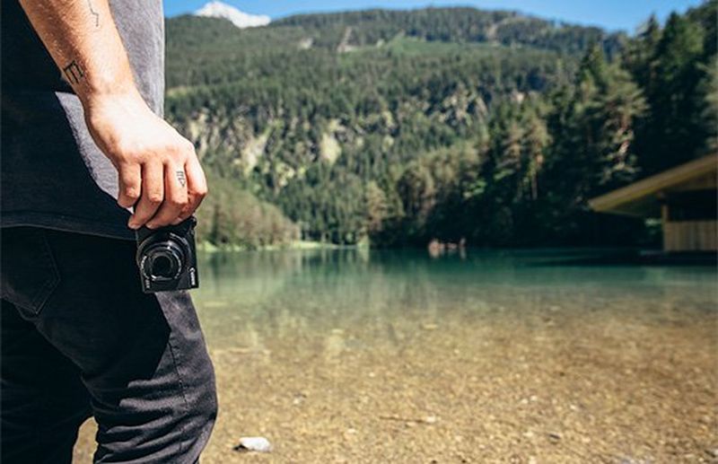 A man, shot from the shoulders down, stands by a lake holding a Canon PowerShot G5 X Mark II in his hand.