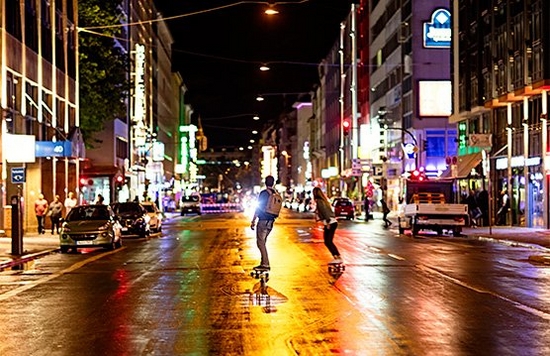 Two people skateboard down a city street at night, colourful lights from buildings reflecting in the wet road surface.