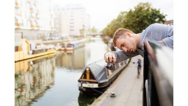 A man leans over some railings to take a picture. Below him is a canal with canal boats moored up.