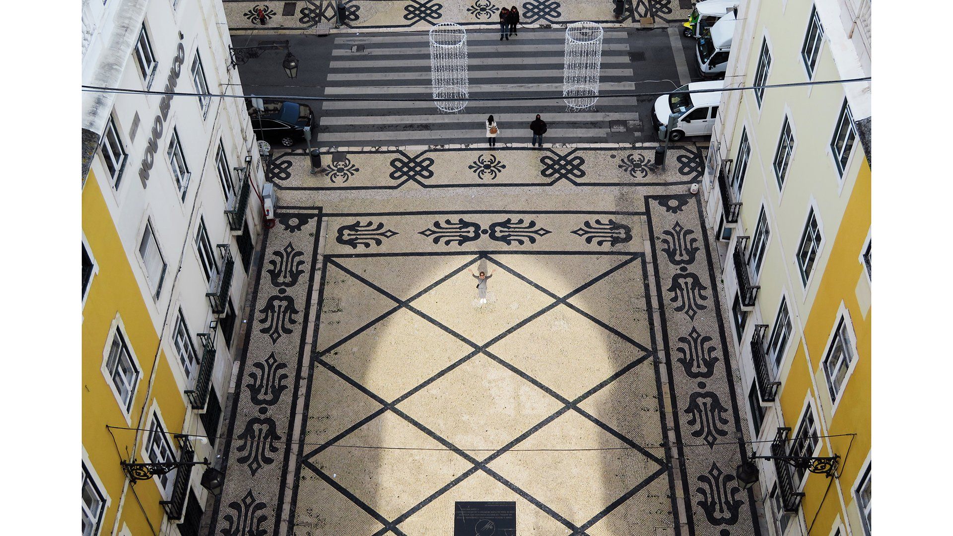 A shot taken from the top of a high building shows a pretty old courtyard with patterned tiled floors and a staircase. On the left and right are yellow and white building fronts.
