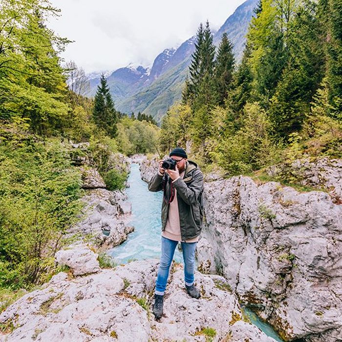 A man stands on rocks in front of a bright blue mountain river, taking a photo on a Canon camera.