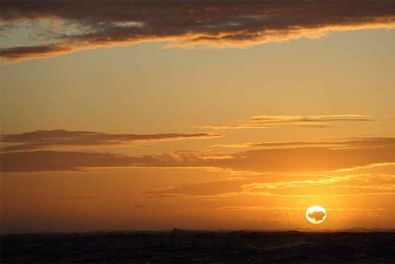The setting sun appears as a disc low on the horizon, illuminating streaky clouds in the orange sky.