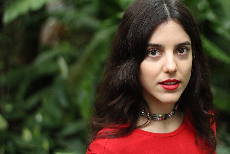 A young woman in sharp focus in the right-hand side of the image, with blurred greenery behind.