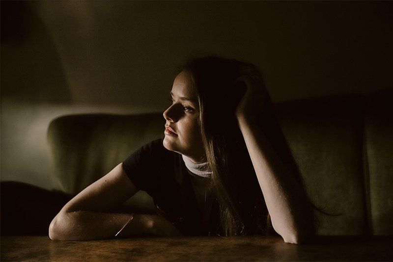 A young woman looking to one side, leaning her head on one hand and the other arm on the tabletop in front of her.