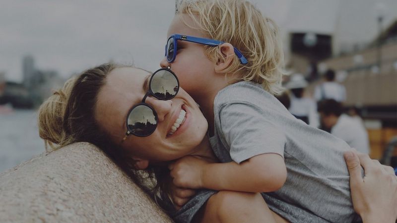A mother picks her son up to hug, both of them wearing sunglasses. Photo by Christian Anderl.