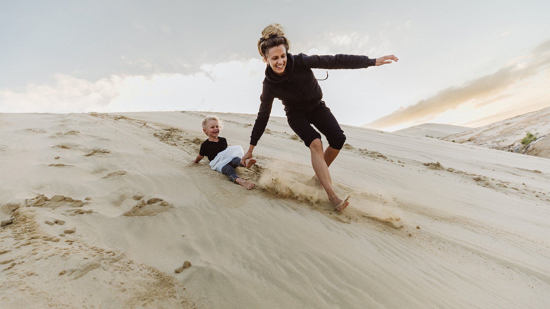 A mother runs down a sand dune, pulling her son along, both of them laughing. Photo by Christian Anderl.
