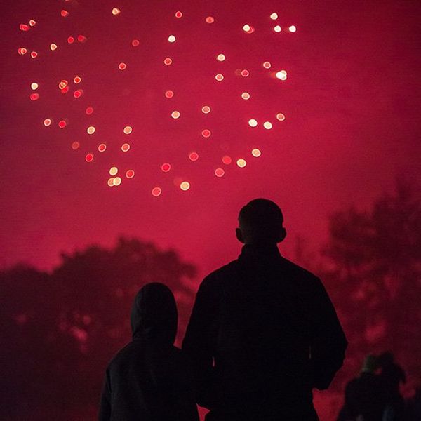 A firework turns the night sky red as it explodes. Below, trees and a crowd of people appear as silhouettes.