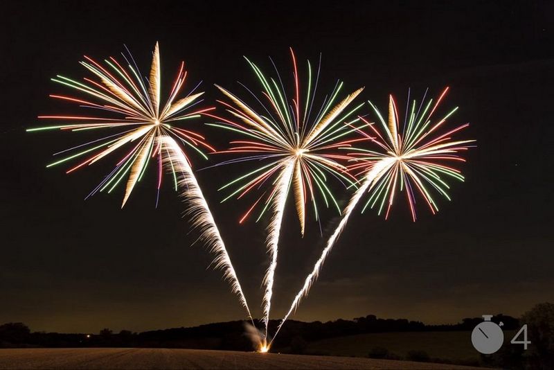 Light trails show the path of three fireworks shooting upwards and bursting in gold, green, blue and red shapes above a field.