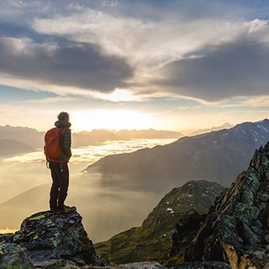 A hiker stands on top of a tall hill or mountain at dawn, arms outstretched.