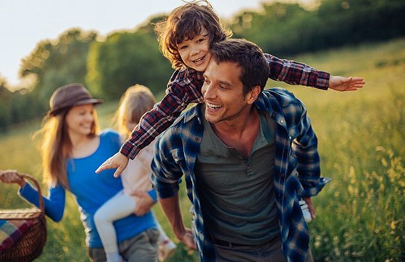 A child on rides on a man's back and a woman carries another child through a sunny field.