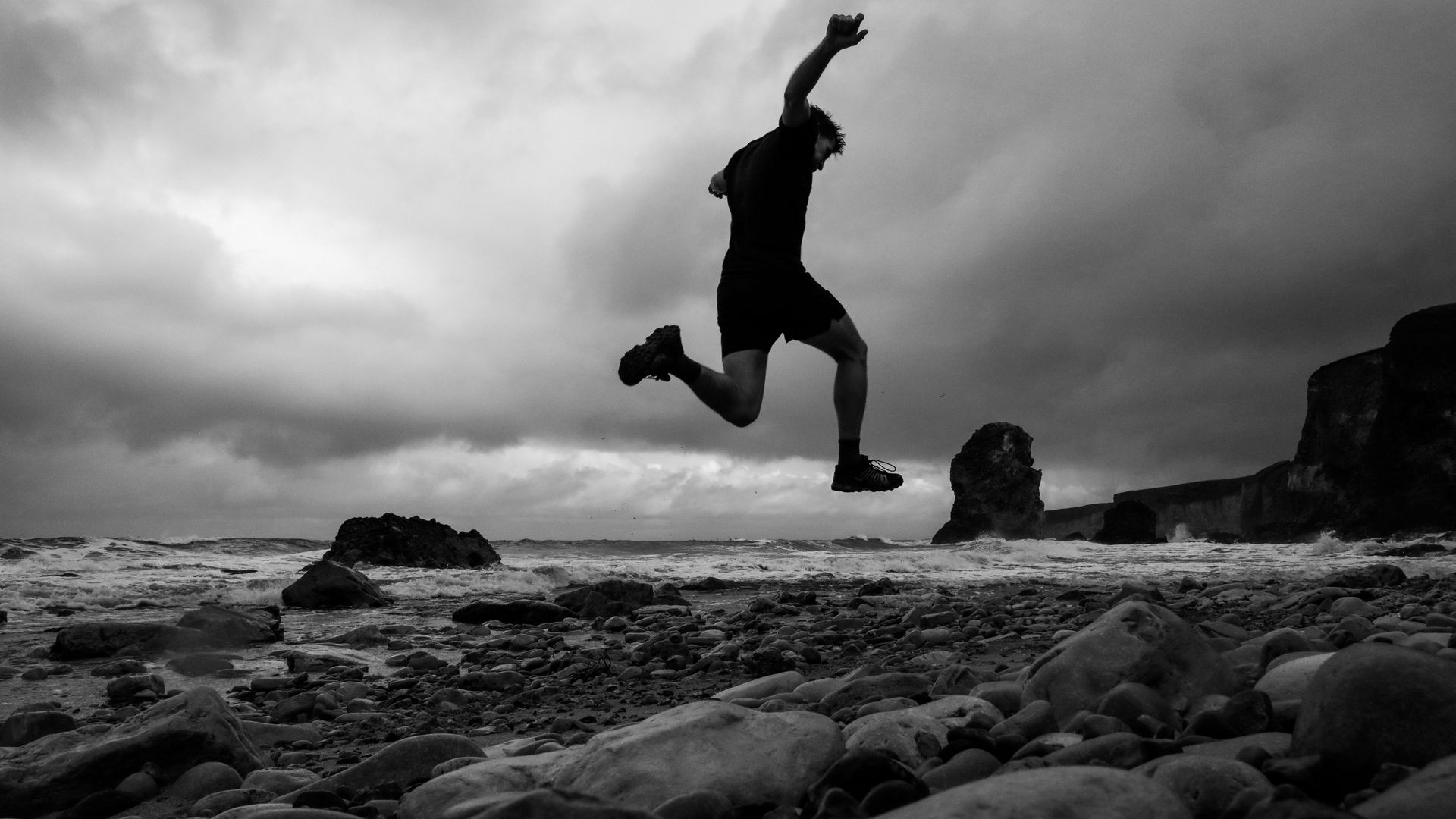 A low-angle shot shows a man in the air as he runs on a rocky beach.