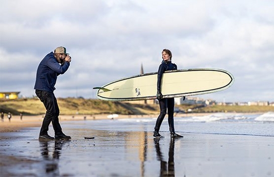 Photographer Tom Bing on a beach taking a picture of a surfer holding her board.