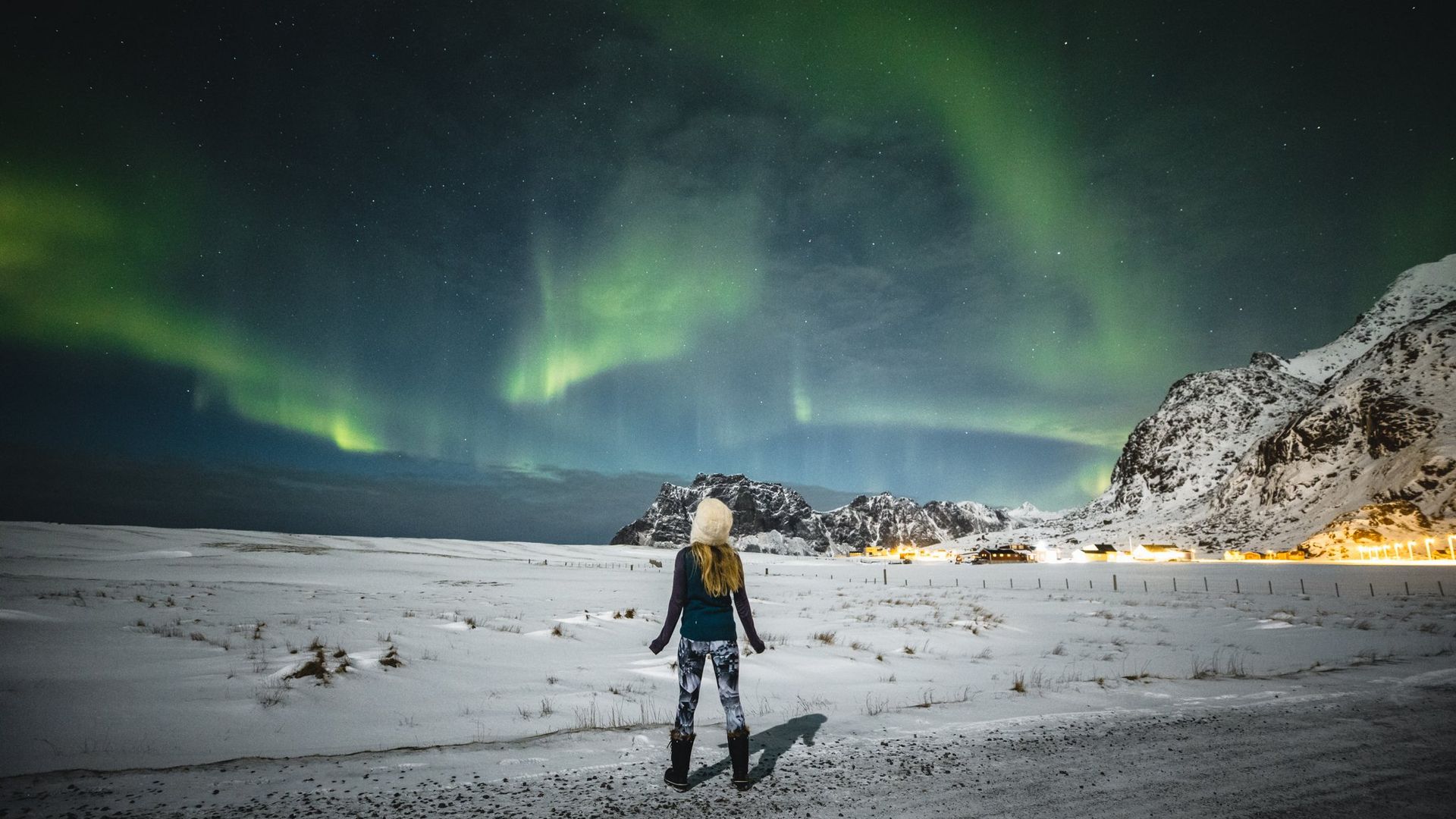 A woman stands on the pale sands of a beach in Norway looking up at the Northern Lights.