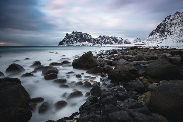 A long exposure shot of water surrounding rocks with a snowy mountain on the horizon.