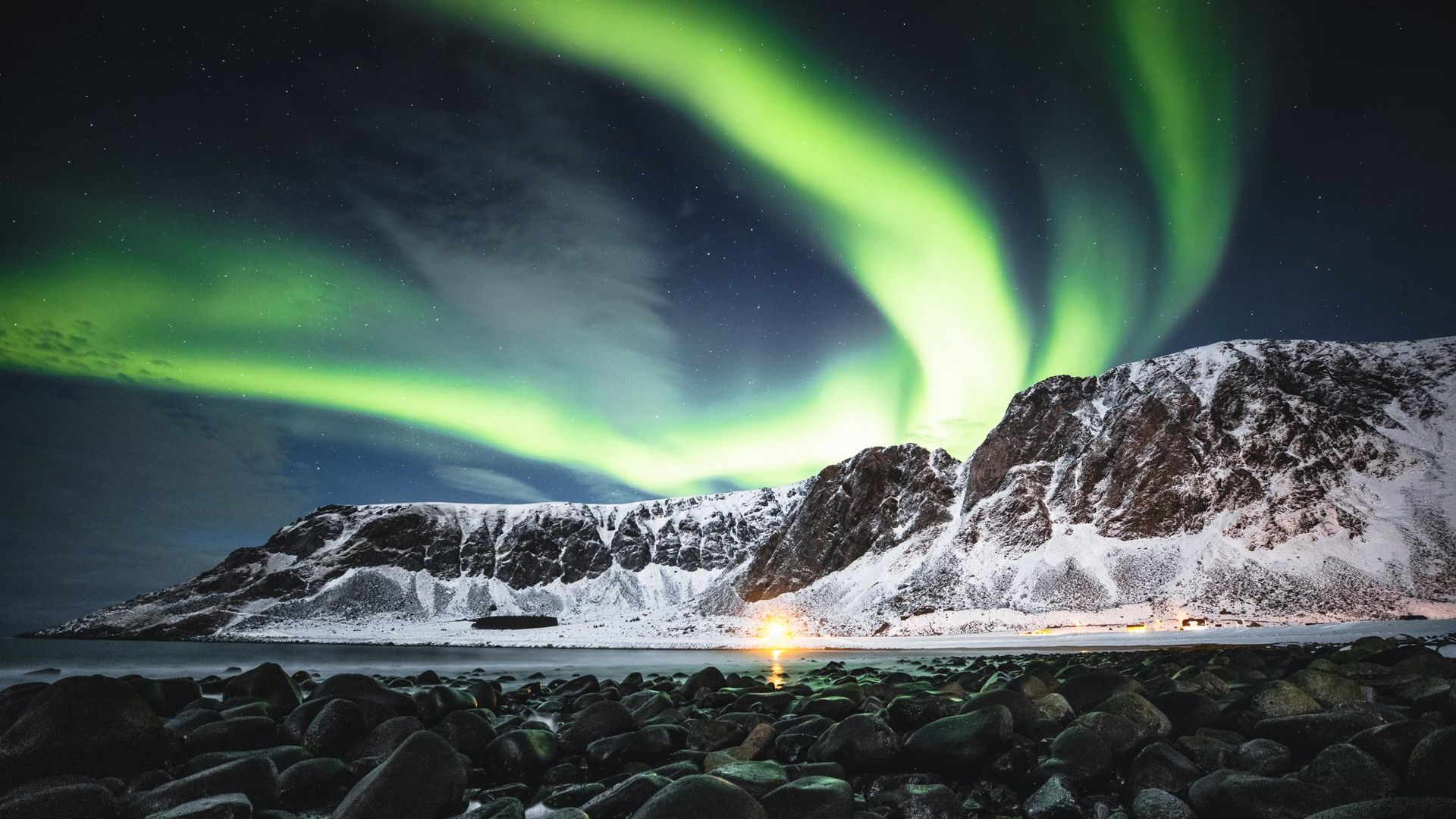 The Northern lights shine brightly across the mountains and the beach below.