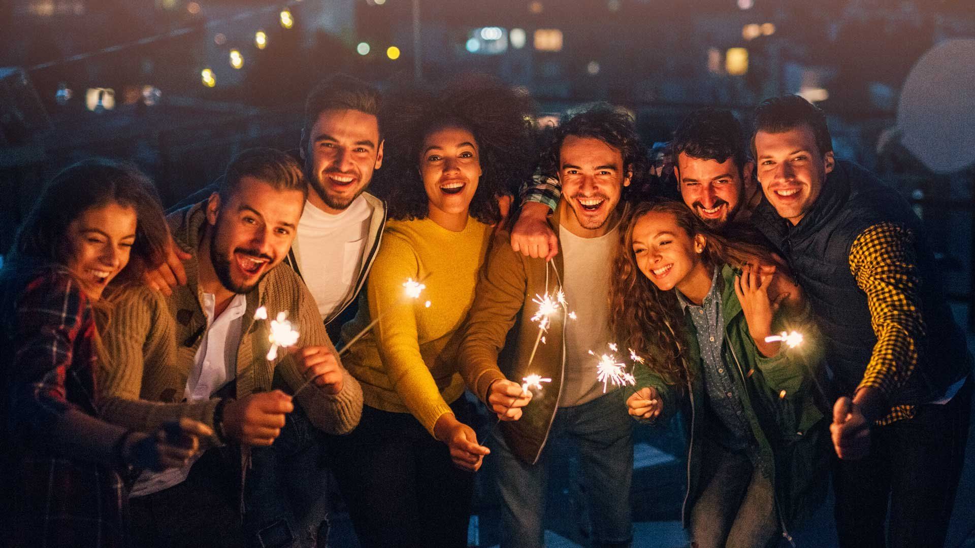 A group of friends pose holding sparklers, facing the camera with their faces illuminated by the sparkler light.