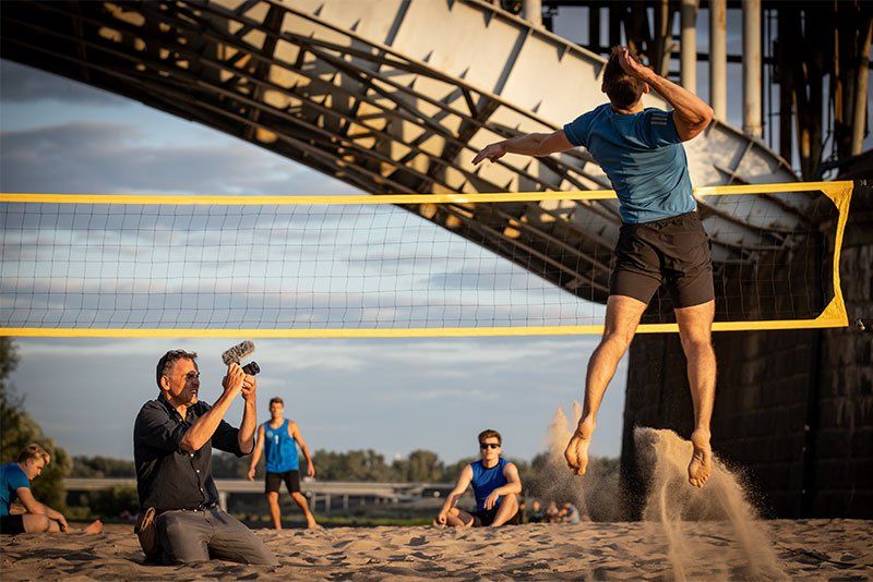 Photographer Piotr Malecki films a volleyball player on a sandy riverbank under a bridge.