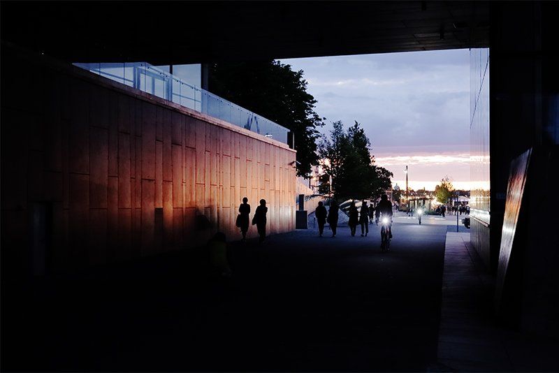 Pedestrians and bike riders on a wide walkway viewed from a dark underpass at sunset.