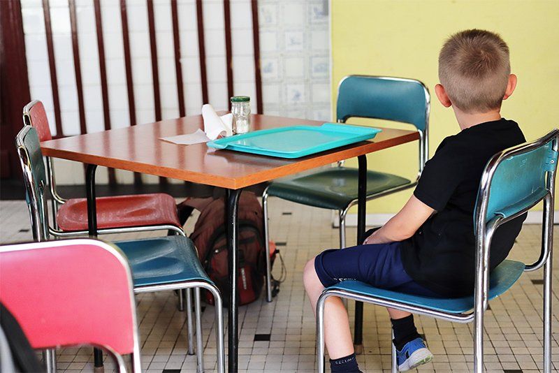 A young boy sits on one of several metal-framed chairs at a table in a cafeteria.