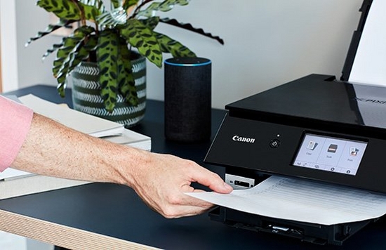 A hand takes a sheet from a printer. An Amazon Alexa is beside the printer.