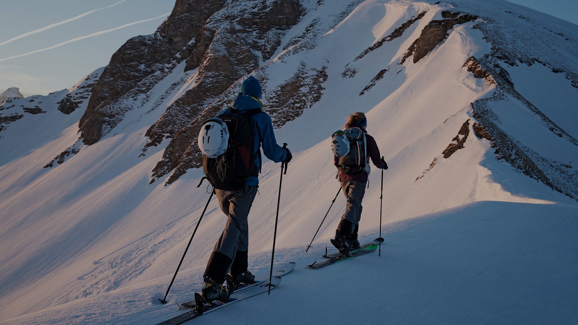Two cross-country skiers travel along the narrow spine of a mountain, using their poles.