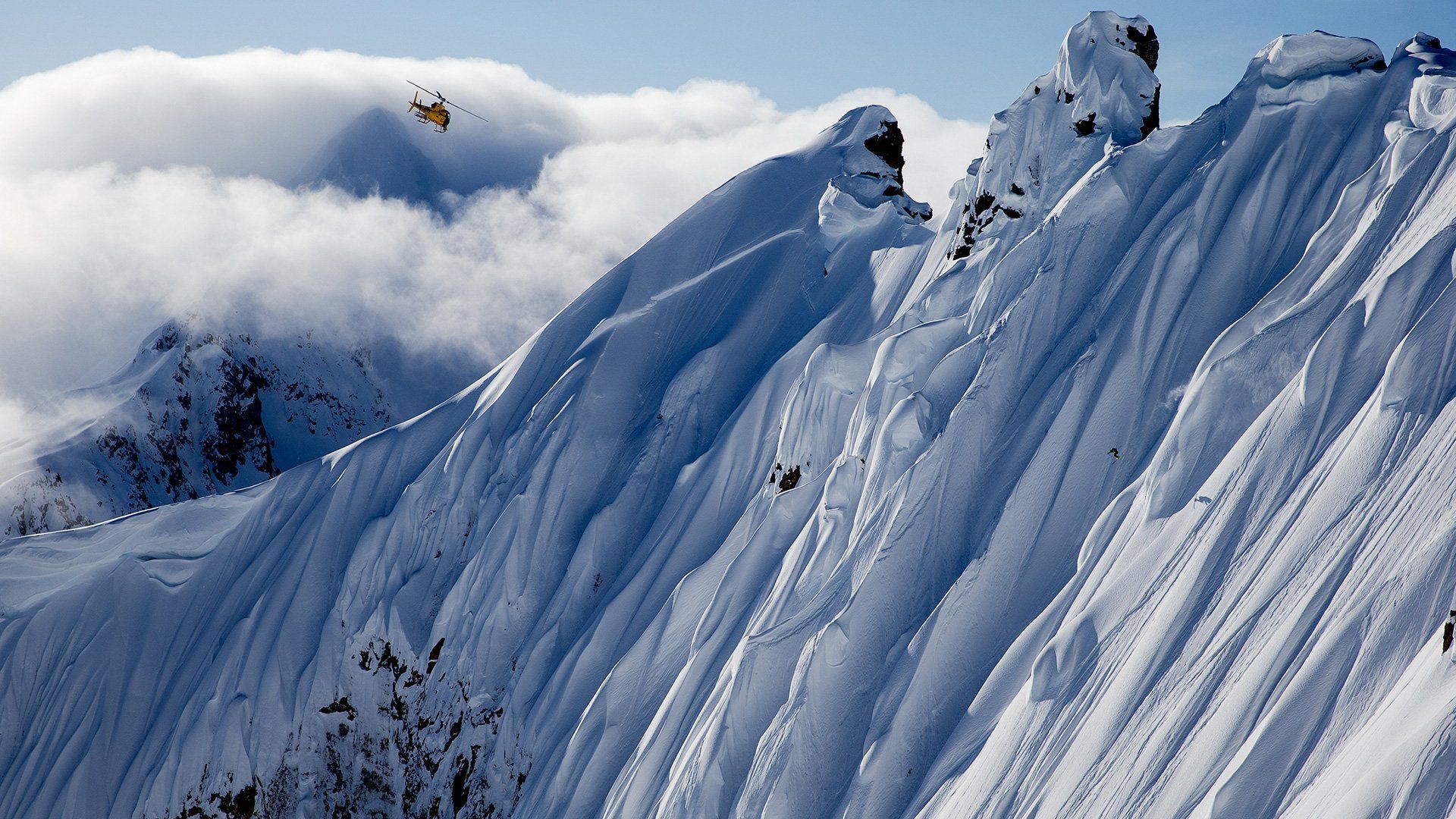 Dwarfed by huge, snow-covered, sheer cliffs near Juneau, Alaska, a member of the Legs of Steel group is barely visible skiing almost vertically downhill as a helicopter hovers above.