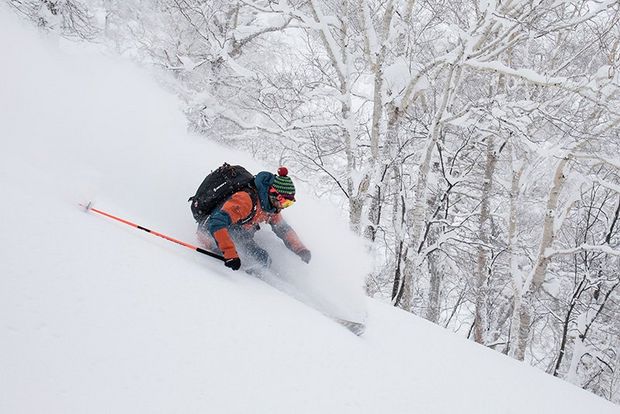 A skier in orange snowsuit and woolly hat sends up a cloud of snow as he travels down a slope by some trees.