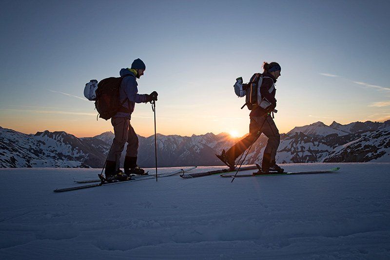 Two cross-country skiers travel along the narrow spine of a mountain, using their poles.