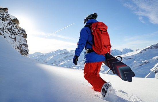 A snowboarder walks through the snow with his snowboard under his arm, away from the camera.