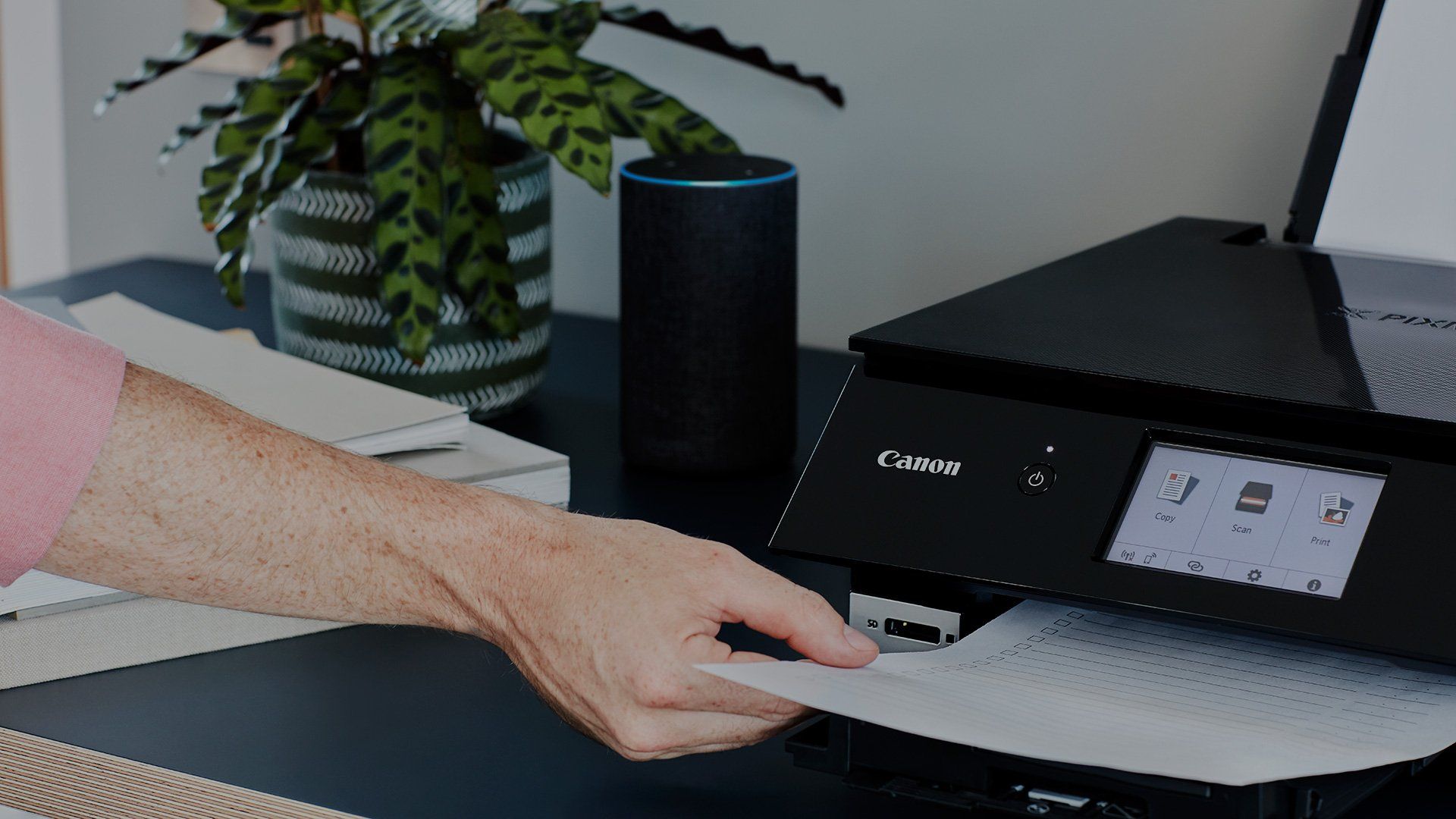 A user takes hold of a printout as it emerges from a black Canon PIXMA TS8250 printer, which sits on a tabletop with an Amazon Echo smart speaker next to it.