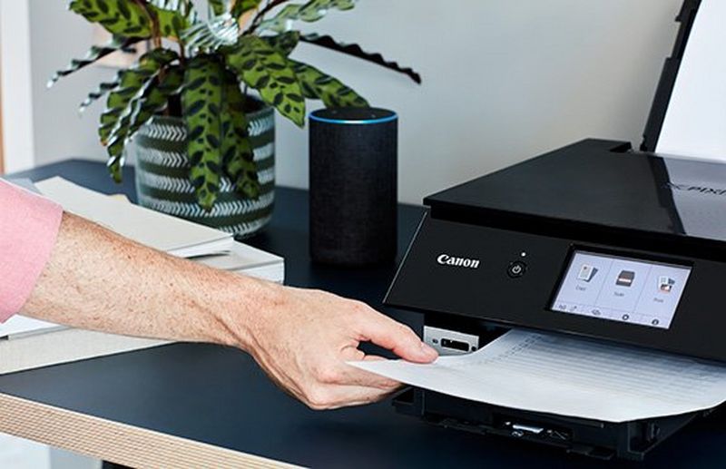 A user takes hold of a printout as it emerges from a black Canon PIXMA TS8250 printer, which sits on a tabletop with an Amazon Echo smart speaker next to it. 