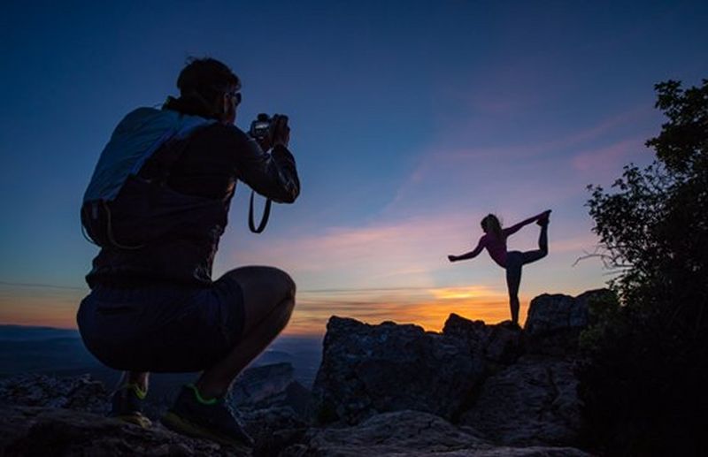 A man squats to photograph a woman standing on one leg, both silhouetted against a sunset.