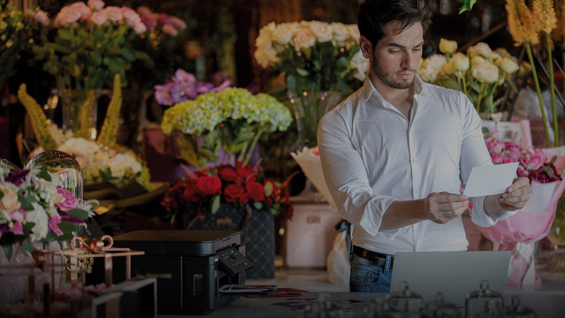 In a room filled with bouquets of flowers, a man holds a print he has made using a Canon PIXMA G6040 that can be seen on the table in front of him.