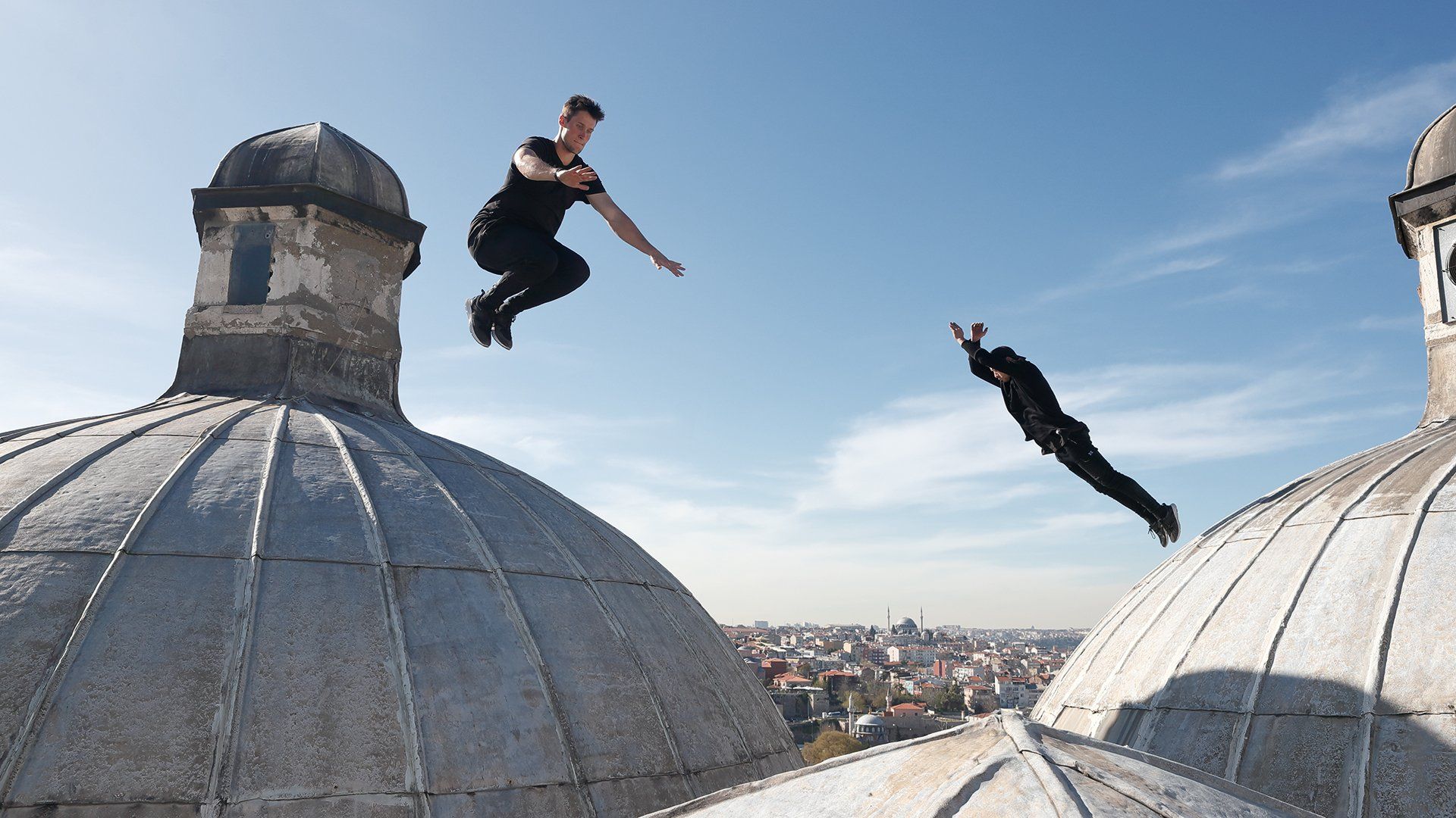 2 young men dressed in black leap around on a rooftop between Turkish domes with stone lanterns on top