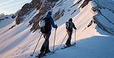 Two cross-country skiers makes their way up a mountain in low sunlight.