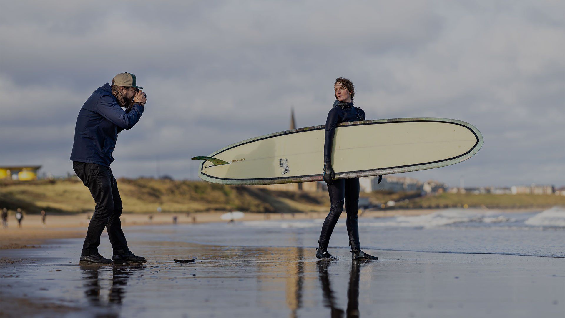 Photographer Tom Bing on a beach taking a picture of a surfer holding her board.