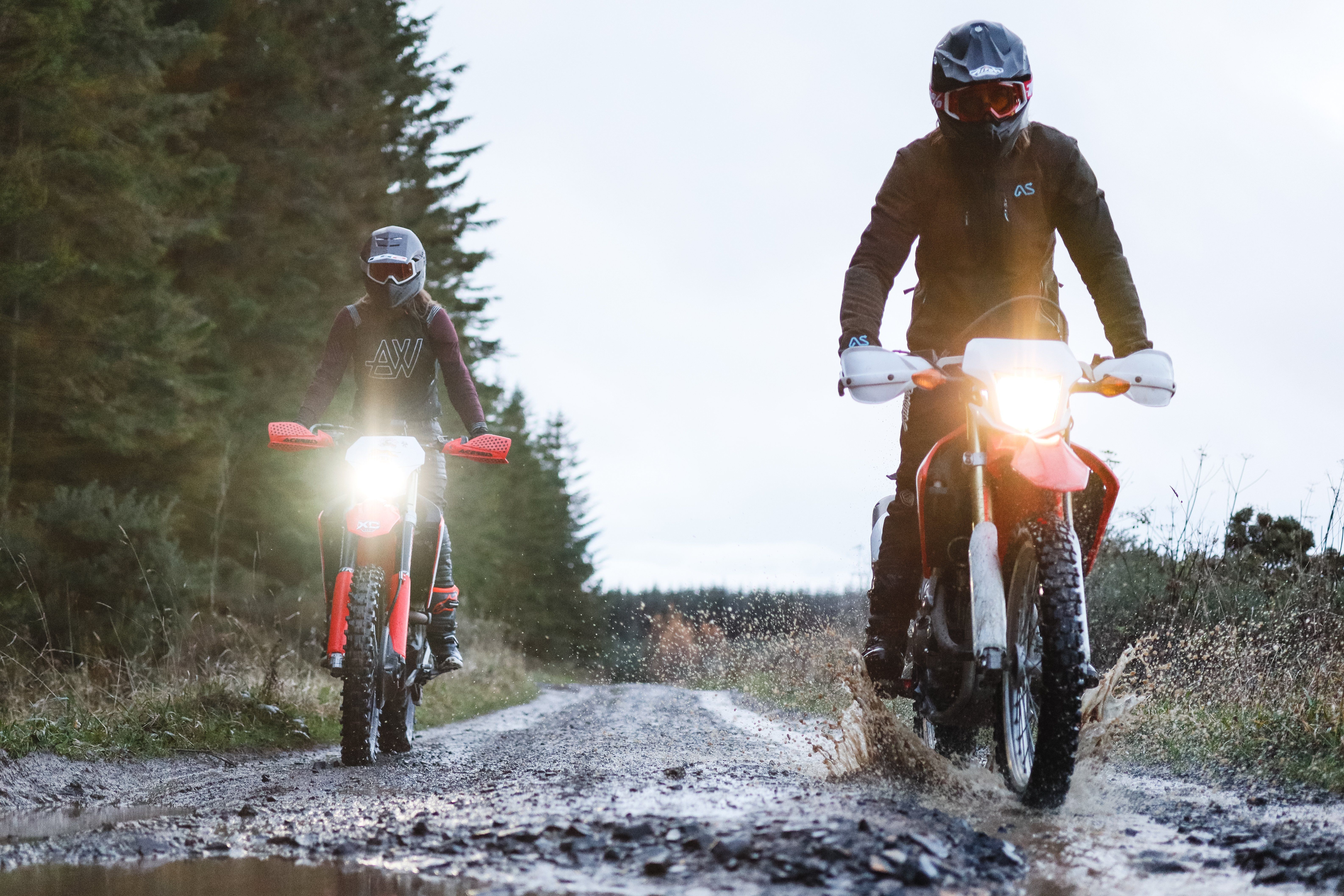 Two riders on dirt bikes approach the camera side-by-side down a dirt track.