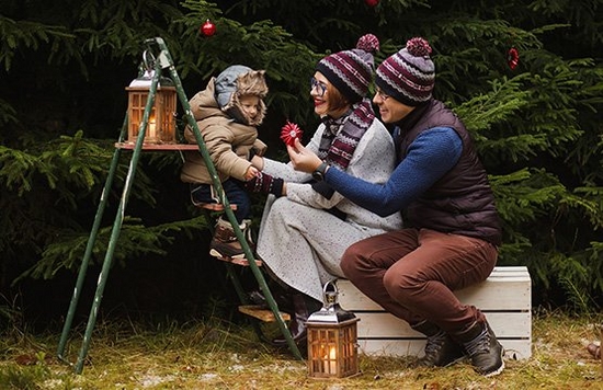 A child in a warm winter coat sits on a stepladder in a garden, entertained by its parents.