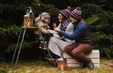 A child in a warm winter coat sits on a stepladder in a garden, entertained by its parents.