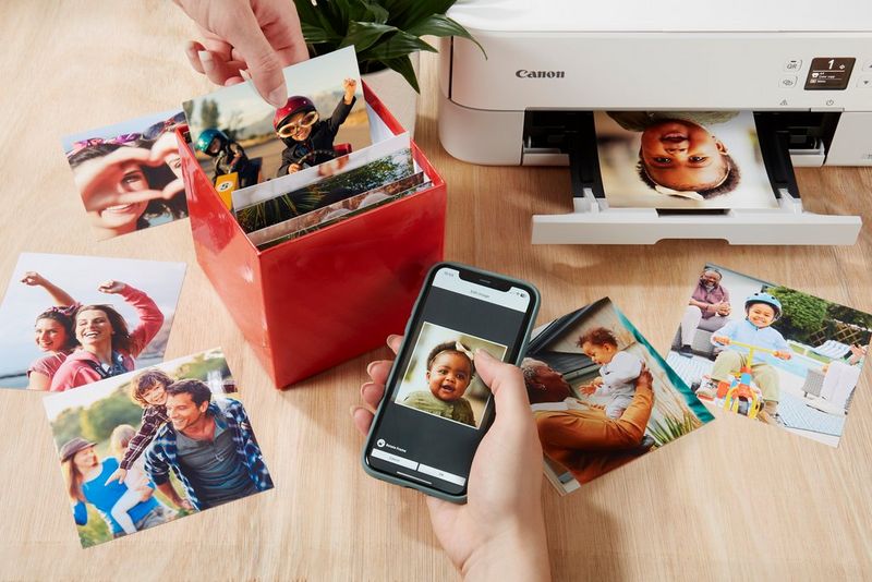 Une main tenant un téléphone avec une photo de bébé à l'écran, tandis qu'une autre main sélectionne une image à partir d'un Canon Photo Cube ouvert.