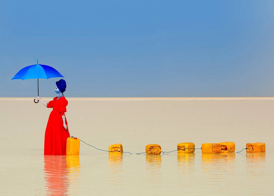 A woman in a red dress and holding a blue umbrella walks through shallow water dragging a chain of jerry cans behind her.