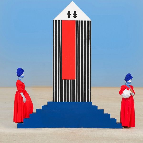 Two women in red dresses and wearing blue headscarves stand either side of a set of blue steps with a black-and-white striped structure on it.