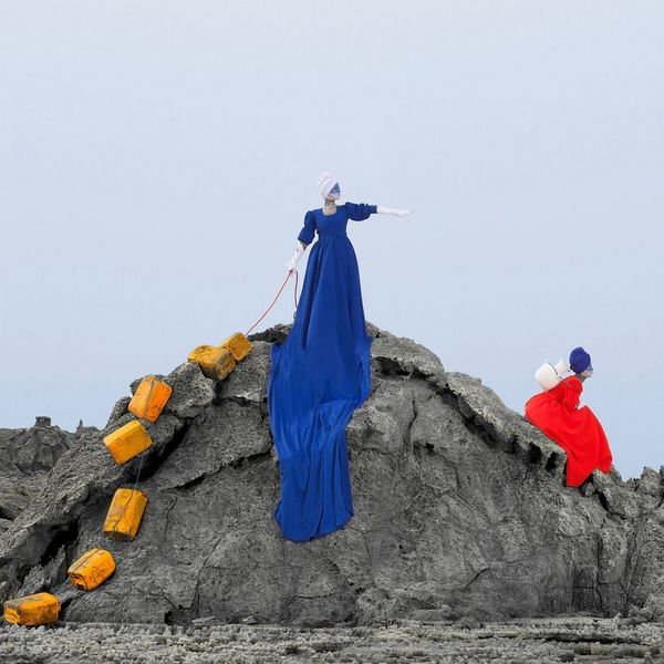 A woman in a long blue dress stands on a rock with a chain of jerry cans behind her. Another woman in a red dress sits on the side of the rock. 